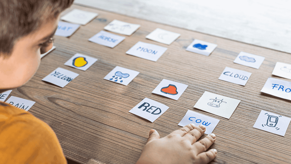 A child playing matching words and pictures on a wooden table. It has text saying yellow, red, and cow.